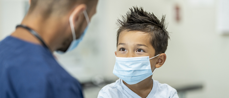 A child wearing a facemask looks up at a doctor during a visit.