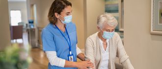 一位戴着口罩的护士微笑着帮助一位老妇人用助行器在养老院里走动。